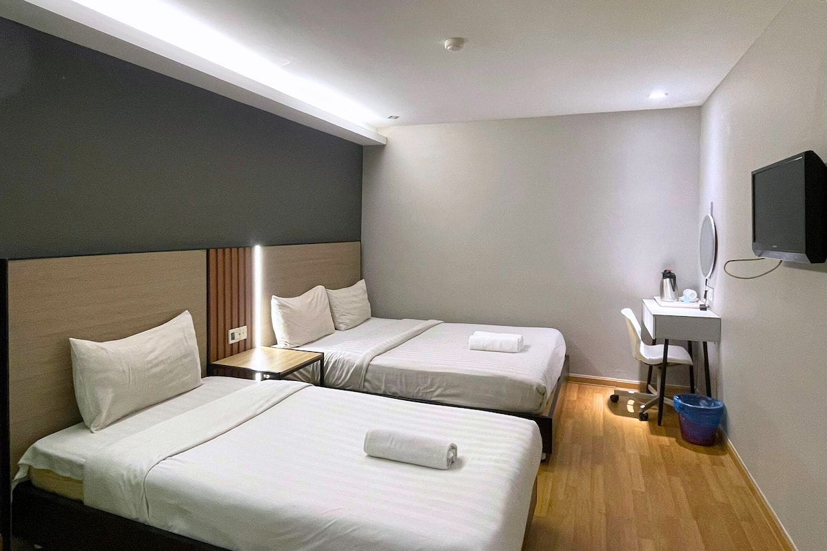 A hotel room with two beds, a desk with a chair, a small table, a TV, and a bathroom mirror.