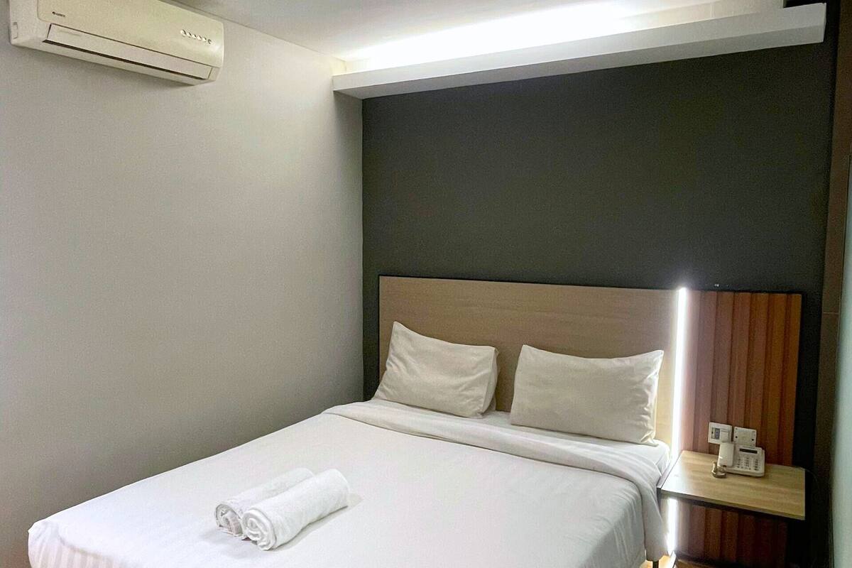 A neatly made bed with white linens and a rolled towel, a bedside table with a phone, and an air conditioning unit on the wall.
