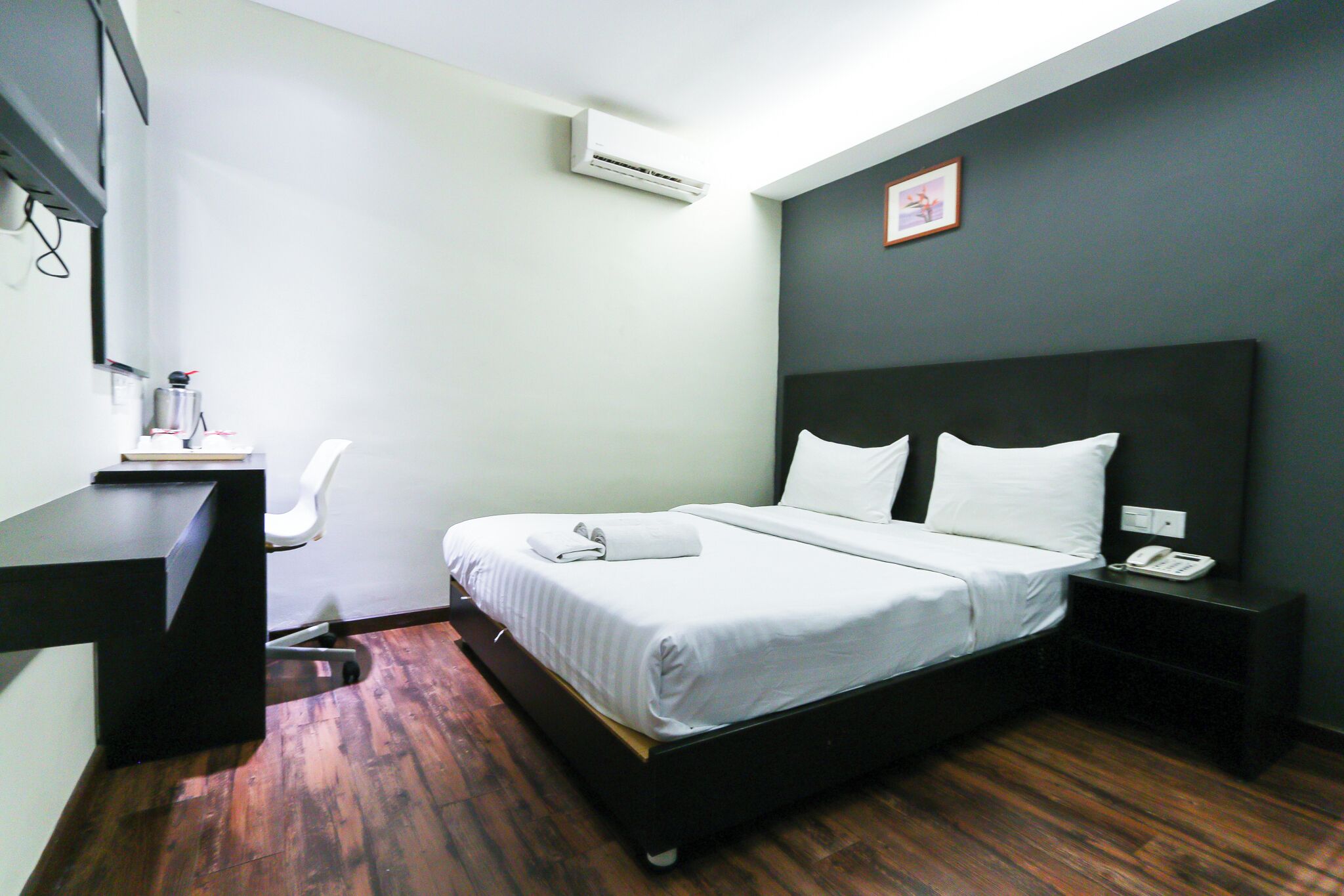 A hotel room with a bed, a desk, a chair, a nightstand, and an air conditioning unit.