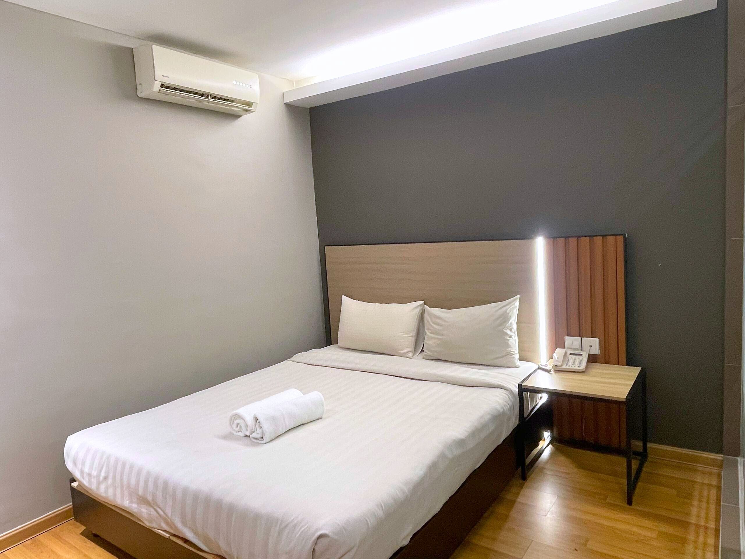 A neatly made bed with white linens and a rolled towel, a bedside table with a phone, and a wall-mounted air conditioner.