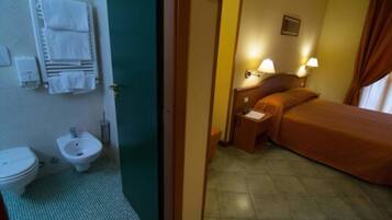 Quarto de hotel com cama, mesa de cabeceira com abajures, pequena mesa com jornal, bidê e porta-toalhas.