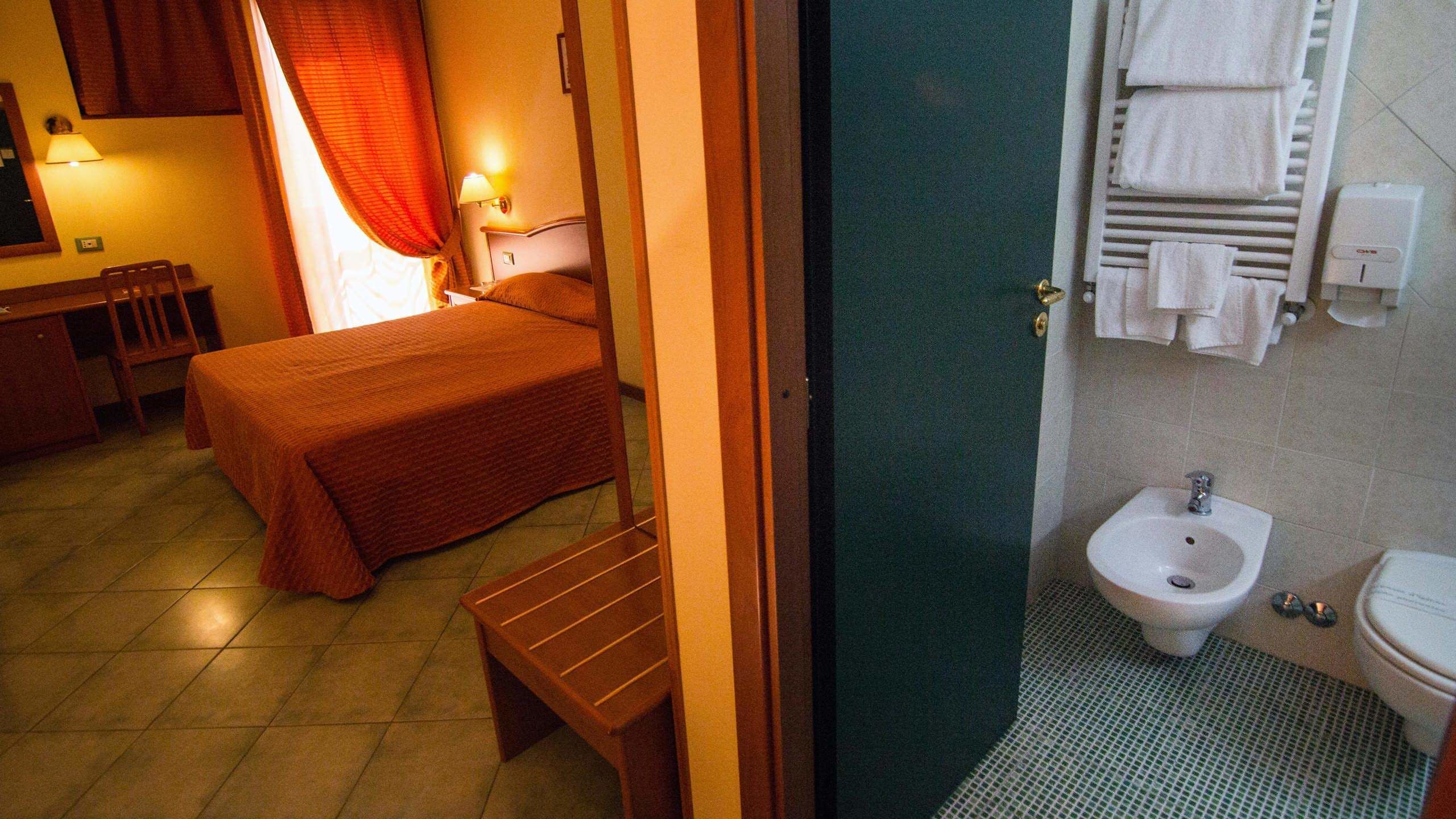 Une chambre d’hôtel comprenant un lit, un bureau, une chaise, une table de chevet, une lampe, une fenêtre avec des rideaux, une salle de bain avec un bidet, des toilettes et un porte-serviettes.