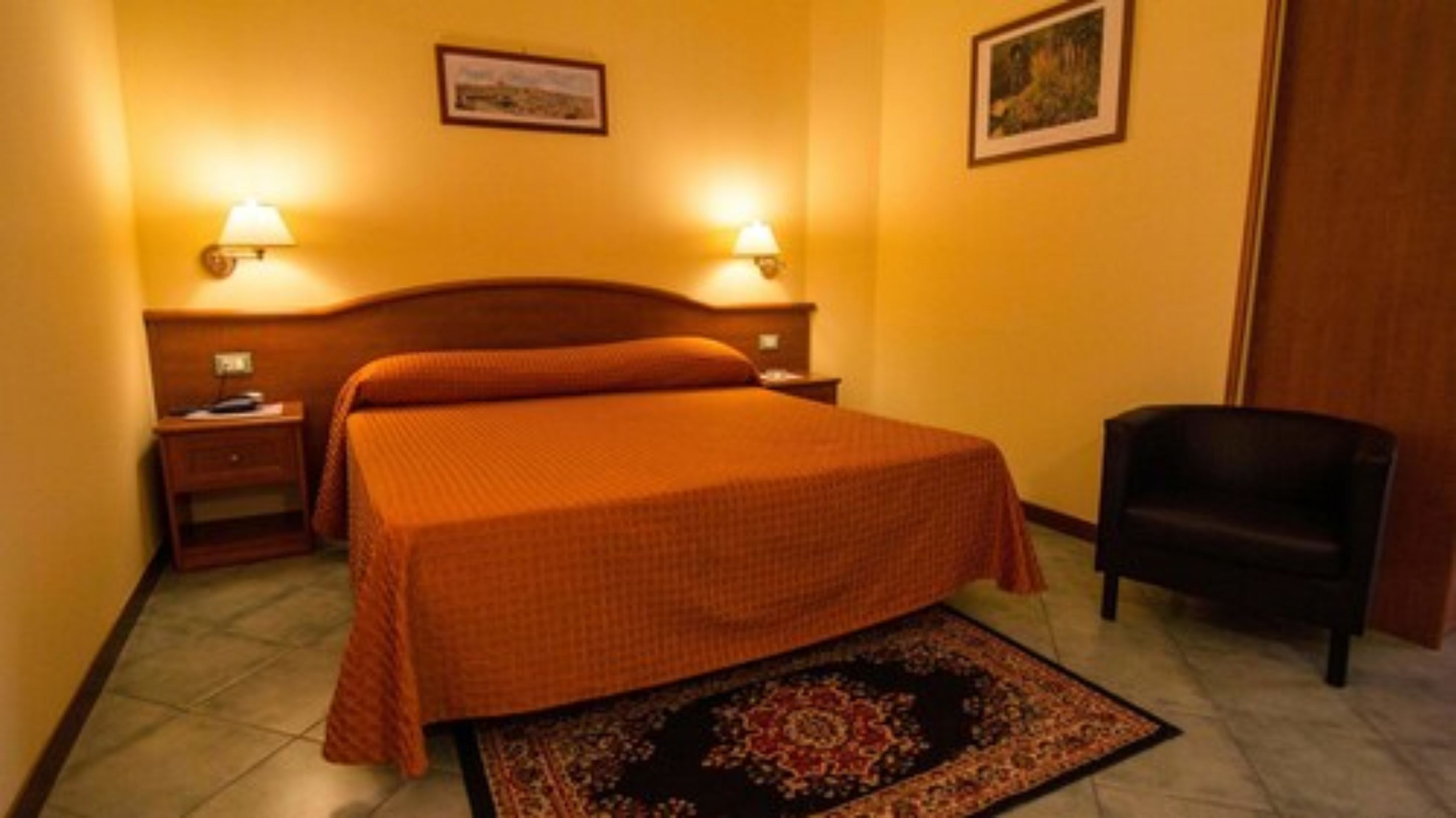 A hotel room with a bed, two bedside tables, two wall-mounted lamps, a chair, and a rug.