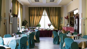 A formal dining room with tables set for a meal, chairs with green covers, and a large window with curtains.