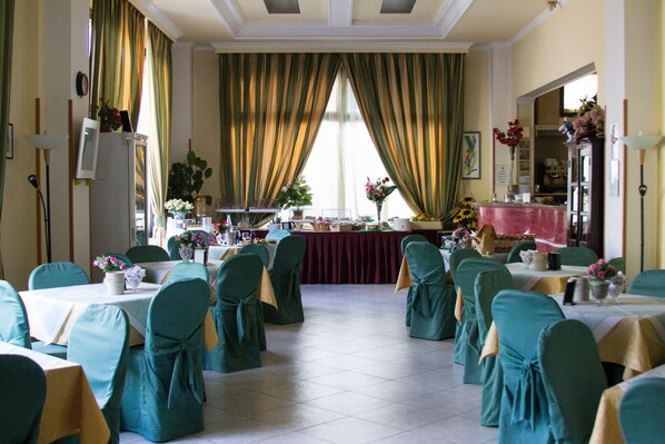 A formal dining room with tables set for a meal, chairs with green covers, and a large window with curtains. - Hotel degli Amici (Artena)