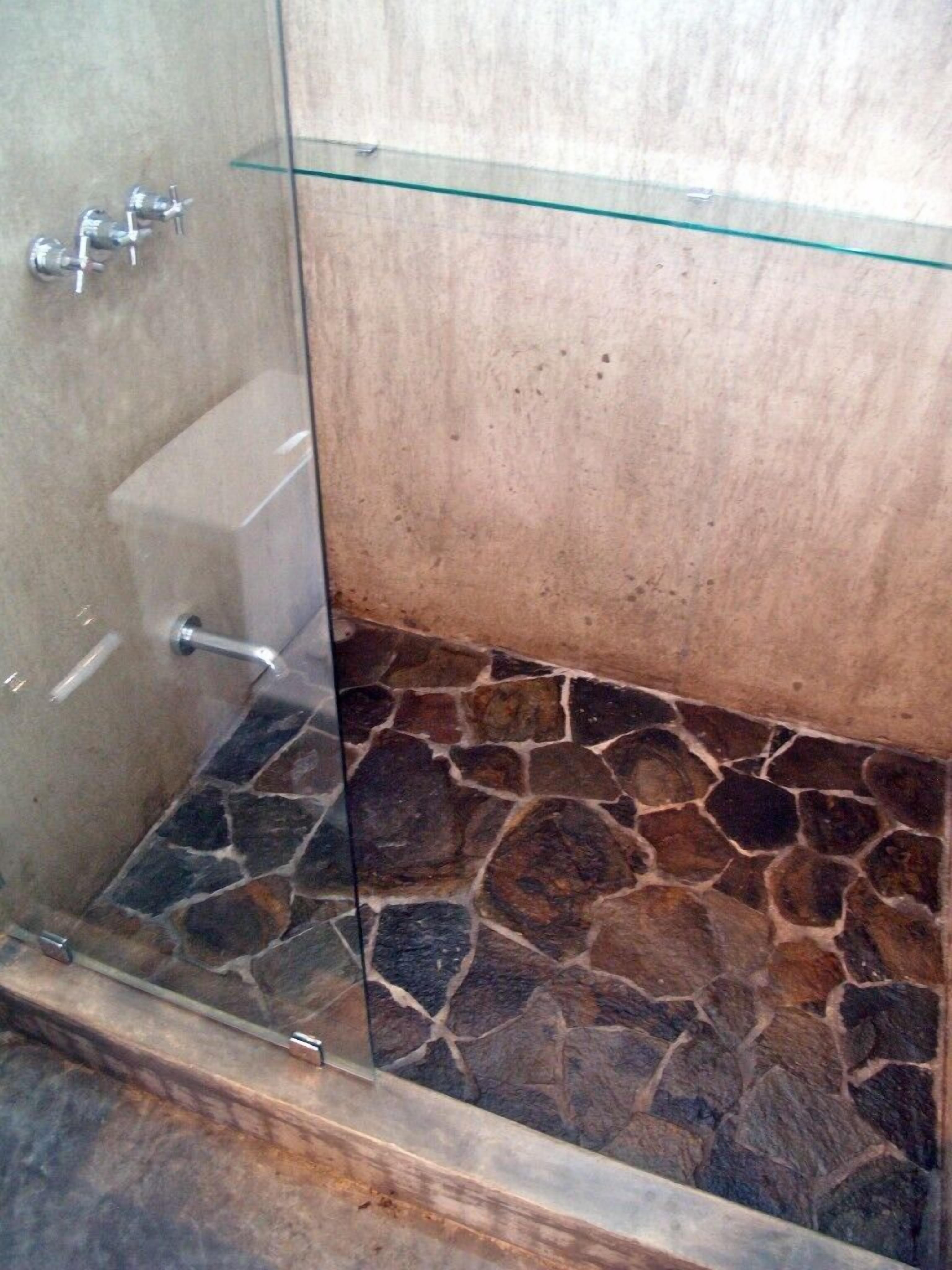 Ducha con mampara de vidrio, suelo de baldosas de piedra y pared color beige.