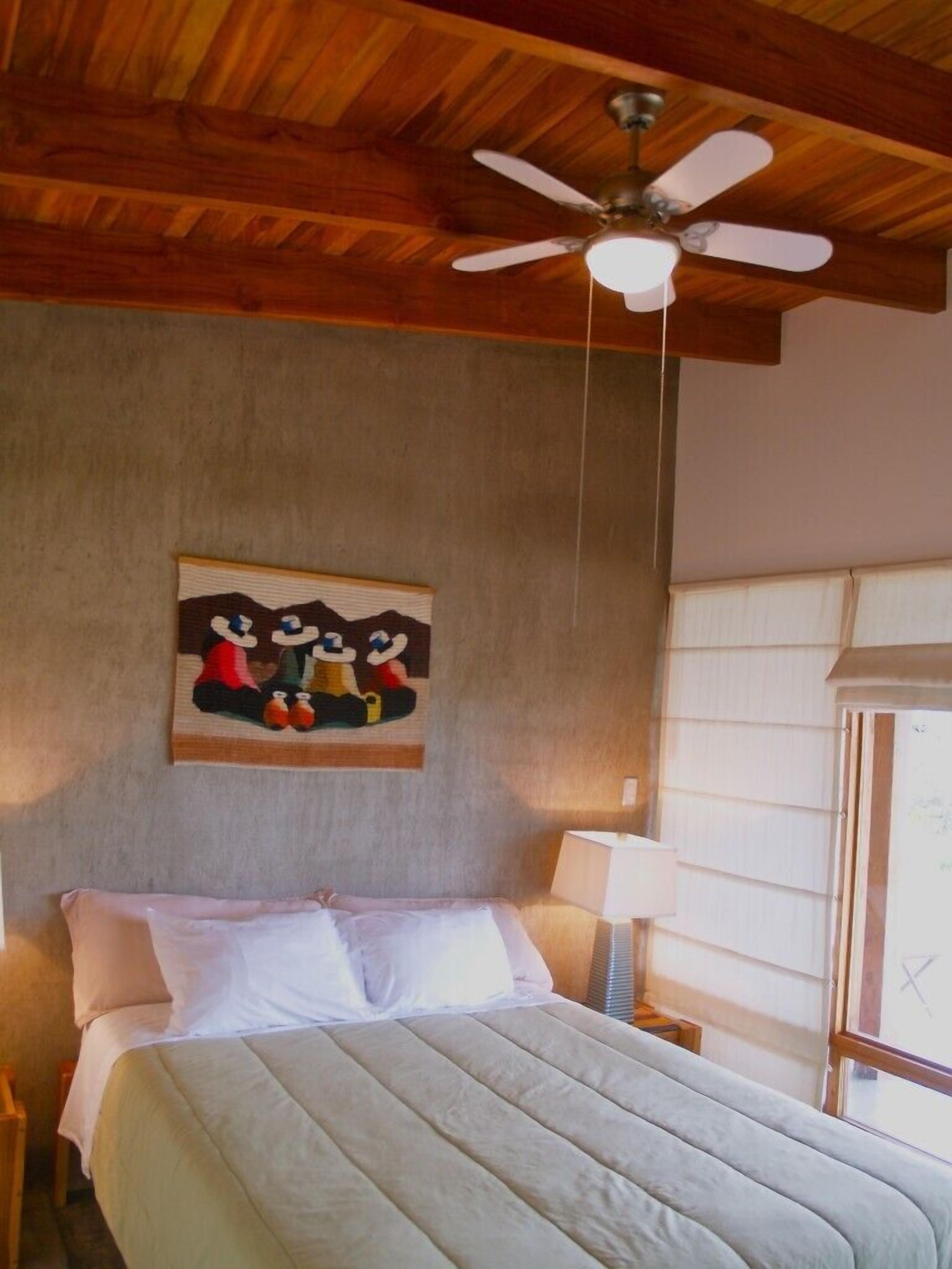 A bedroom with a bed, a ceiling fan, a painting, a window with blinds, and a nightstand with a lamp.
