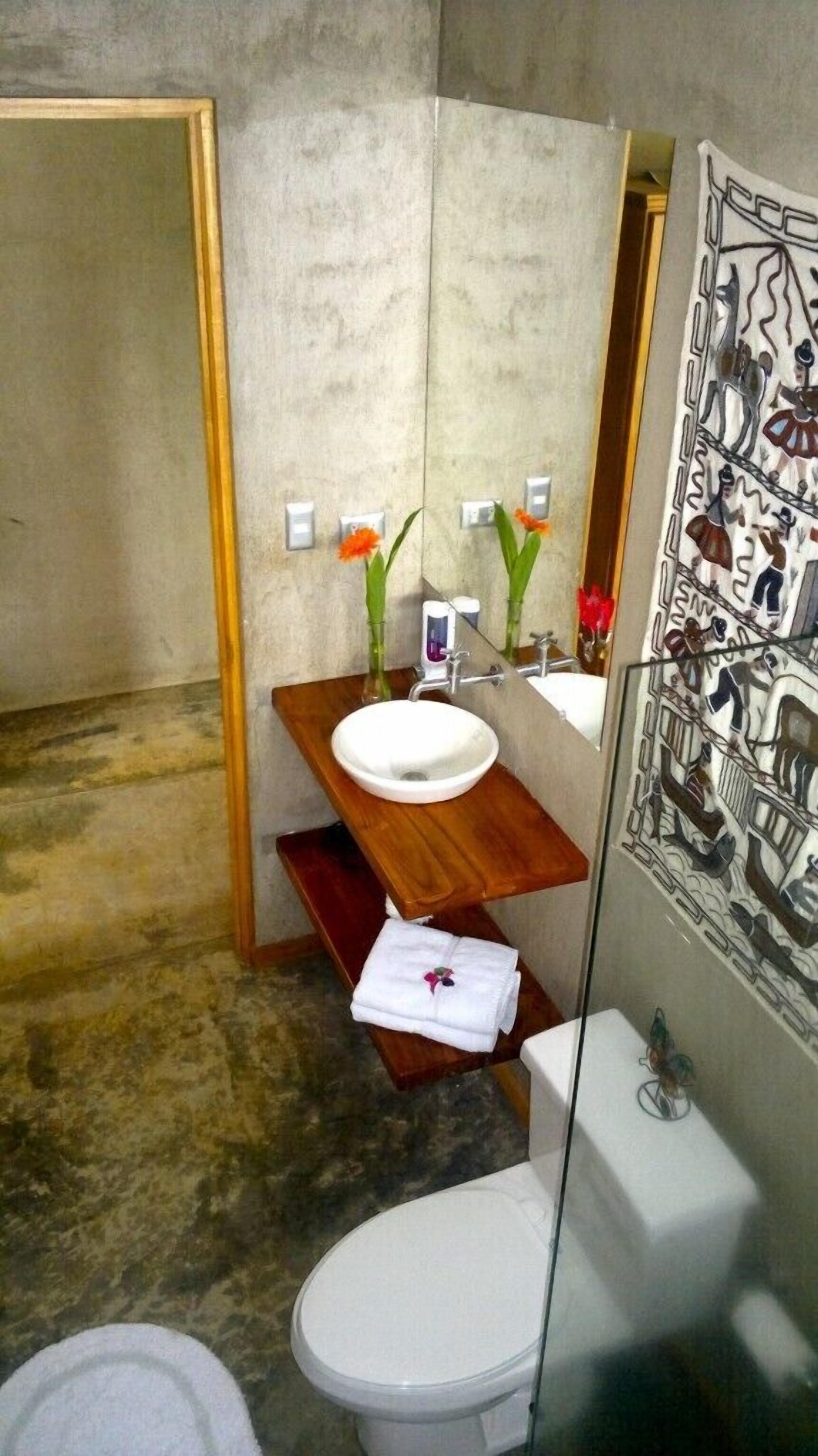 Un baño con tocador de madera, lavabo blanco y retrete. Hay un espejo sobre el lavabo y un tapiz decorativo en la pared.