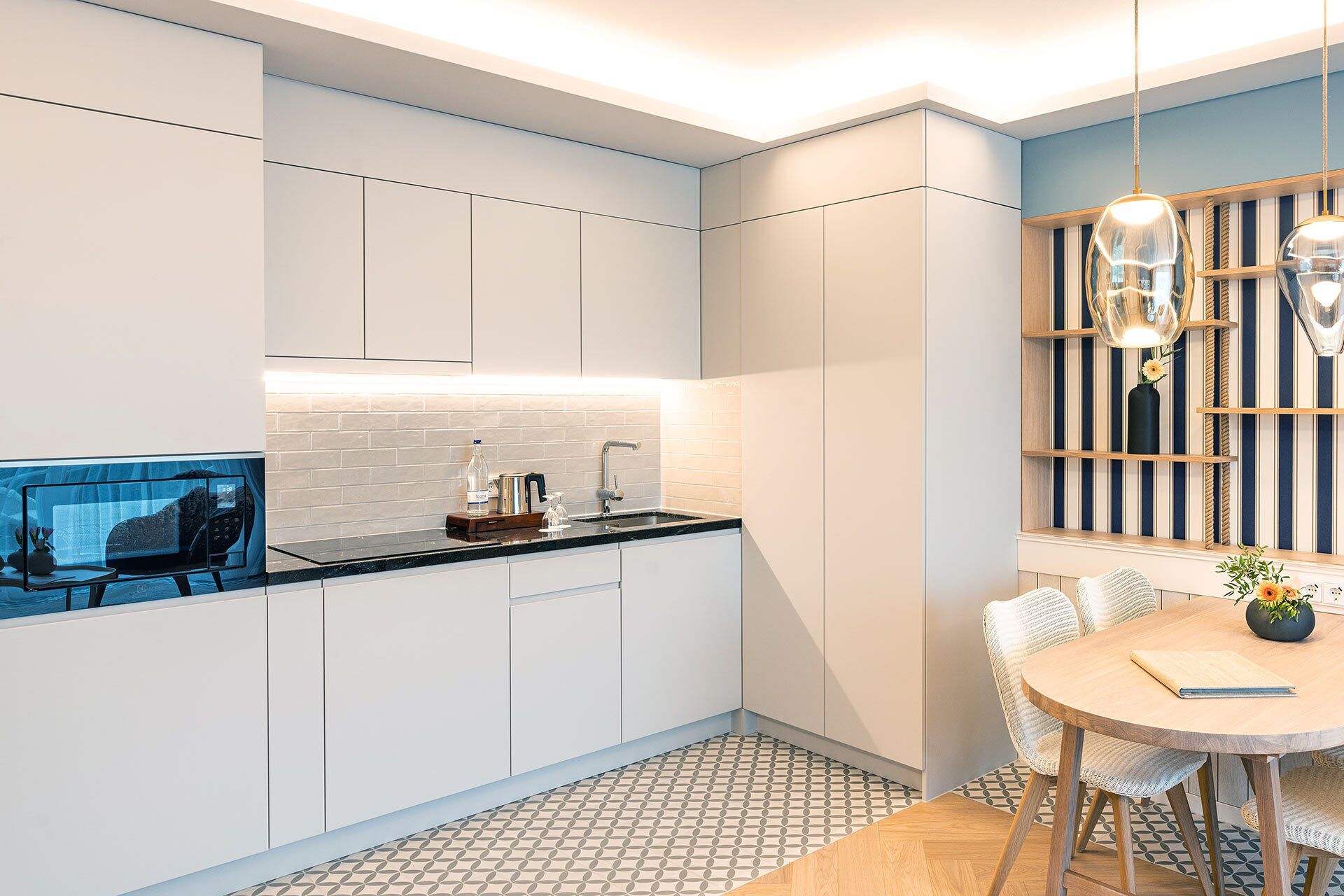 A modern kitchen with white cabinets, a black countertop, and a tiled backsplash. There is a dining area with a round table and chairs.