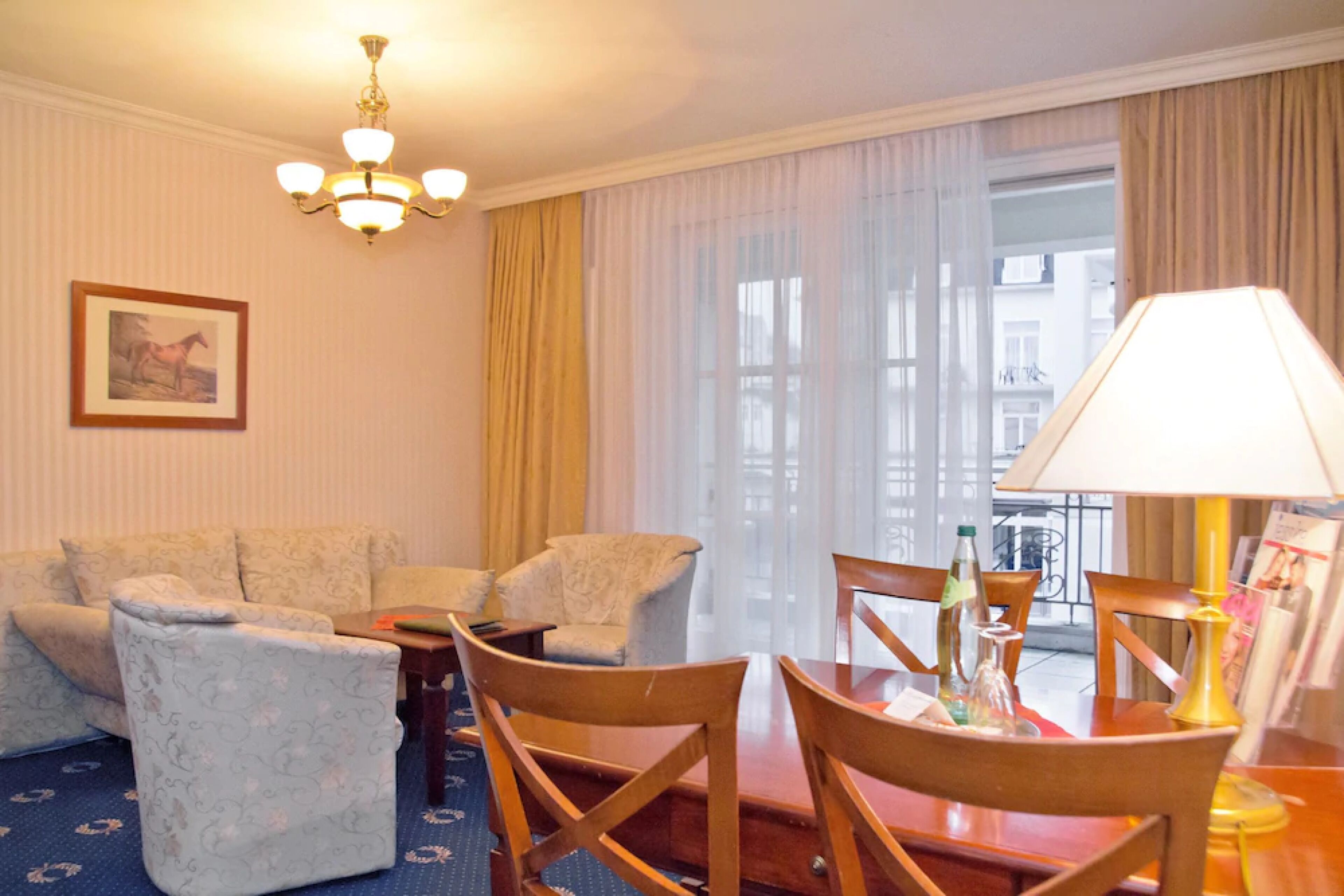 A hotel room with a sofa set, a dining table with chairs, a chandelier, and a large window with curtains.