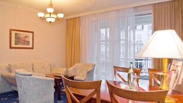 A hotel room with a sofa set, a dining table with chairs, a chandelier, and a large window with curtains.