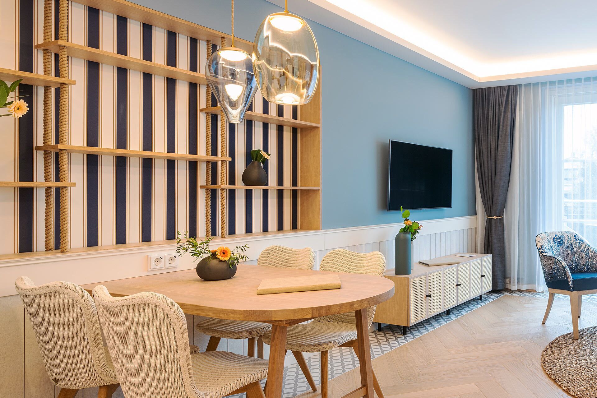 Een moderne woonkamer met een ronde houten eettafel, rieten stoelen, een aan de muur bevestigde televisie en een groot raam met gordijnen.