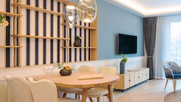 Een moderne woonkamer met een ronde houten eettafel, rieten stoelen, een aan de muur bevestigde televisie en een groot raam met gordijnen.