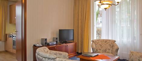 Une chambre d’hôtel équipée d’une télévision, de deux fauteuils à motifs, d’une table en bois, d’une petite armoire et d’une grande fenêtre avec des rideaux.