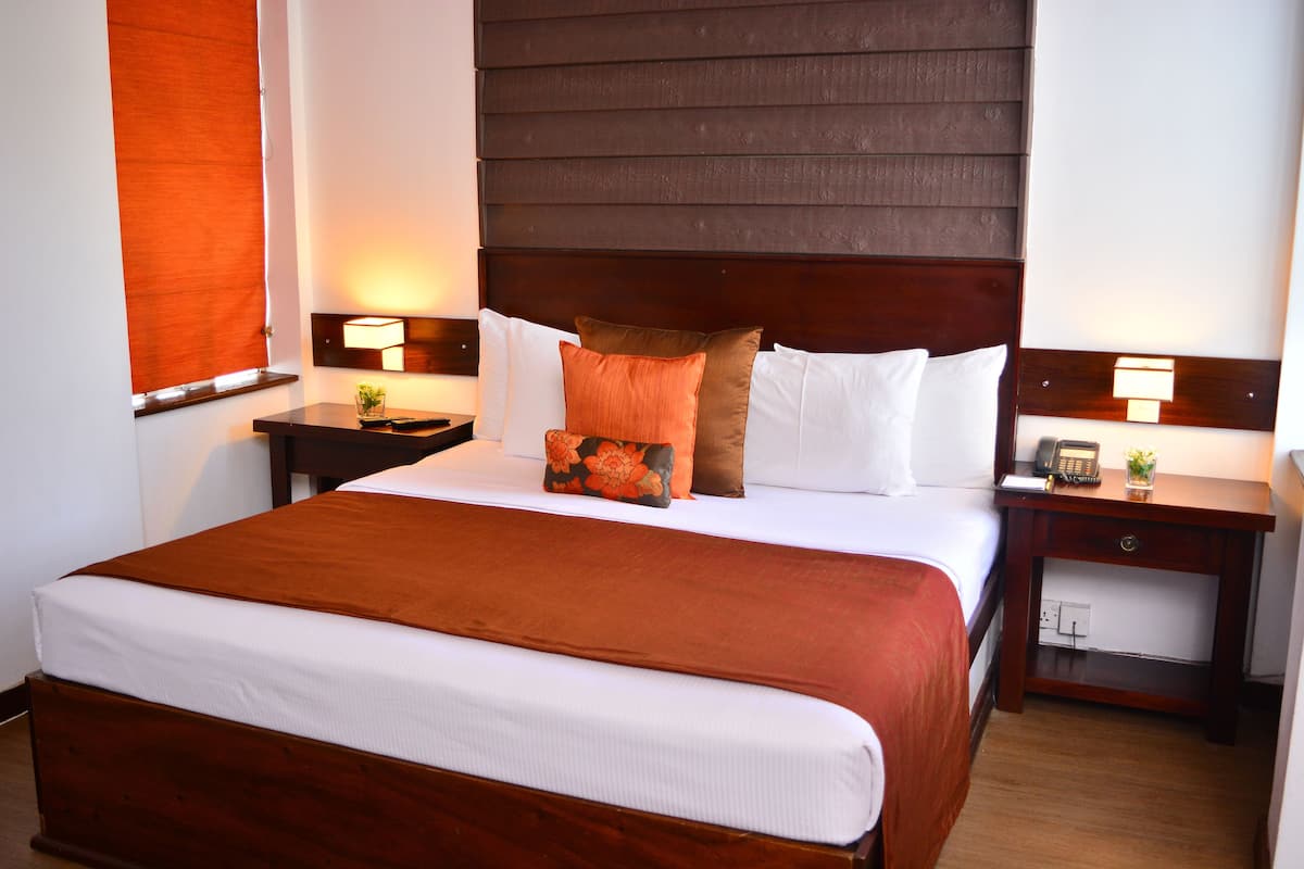 A neatly made bed with a brown headboard, white bedding, and a mix of white and brown pillows. There are two bedside tables with lamps and a small plant on one.