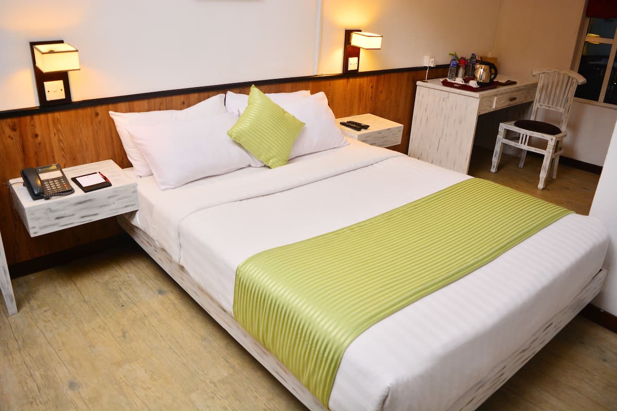 A hotel room with a bed, a nightstand with a phone and remote, a desk with a chair, and a lamp.