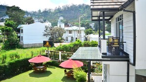 A hotel with a balcony, a garden with two red umbrellas, and a view of the surrounding buildings. - Unique Cottages (Nuwara Eliya)