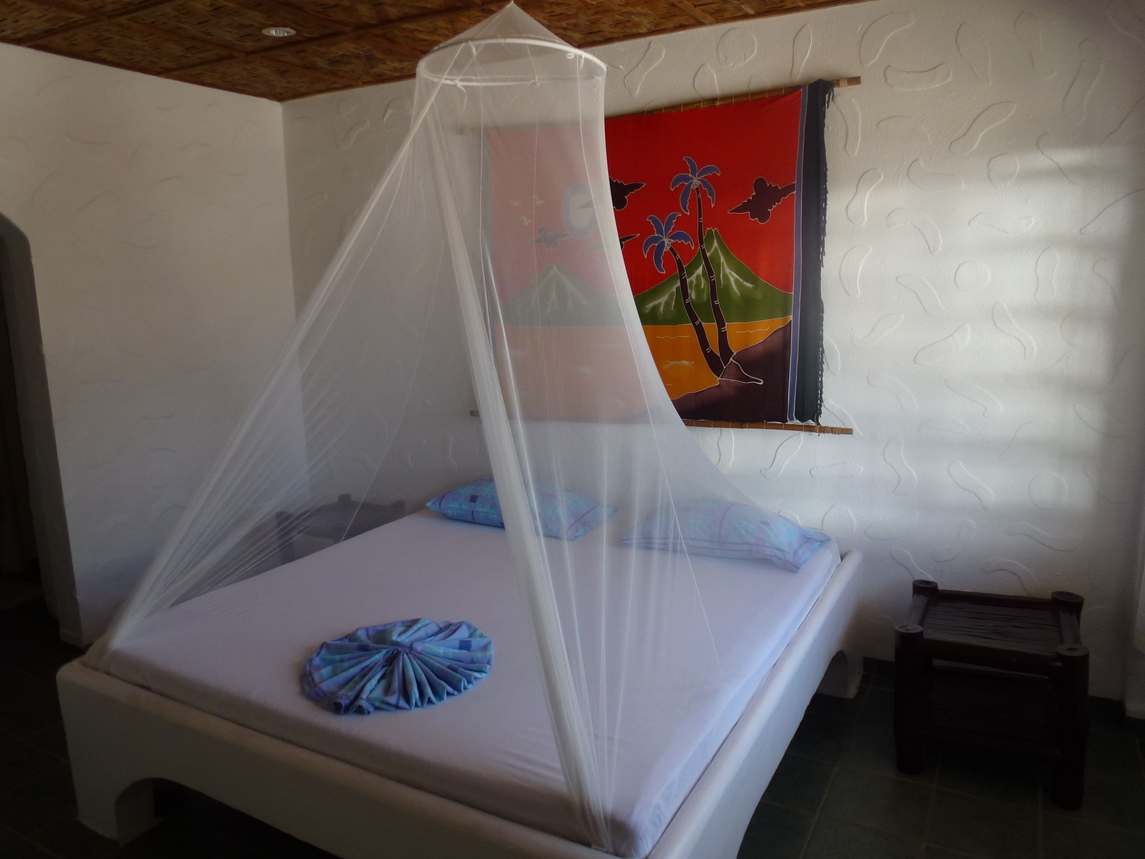 A bed with a mosquito net, a blue pillow, and a painting of palm trees on the wall.