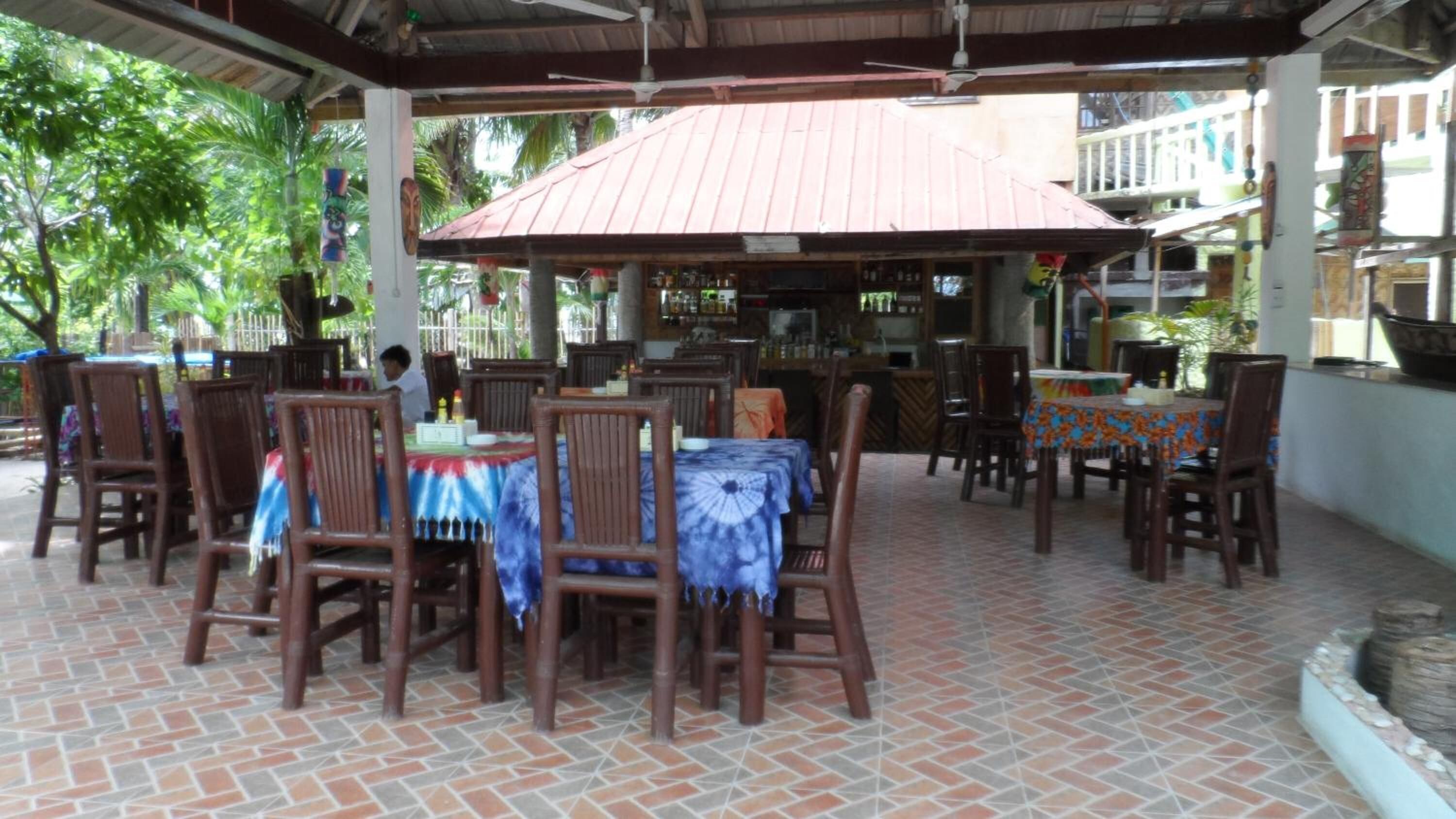 An outdoor restaurant with wooden tables and chairs, a tiled floor, and a bar area in the background.