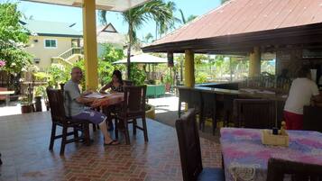 An outdoor restaurant with wooden tables and chairs, a tiled floor, and a bar area.