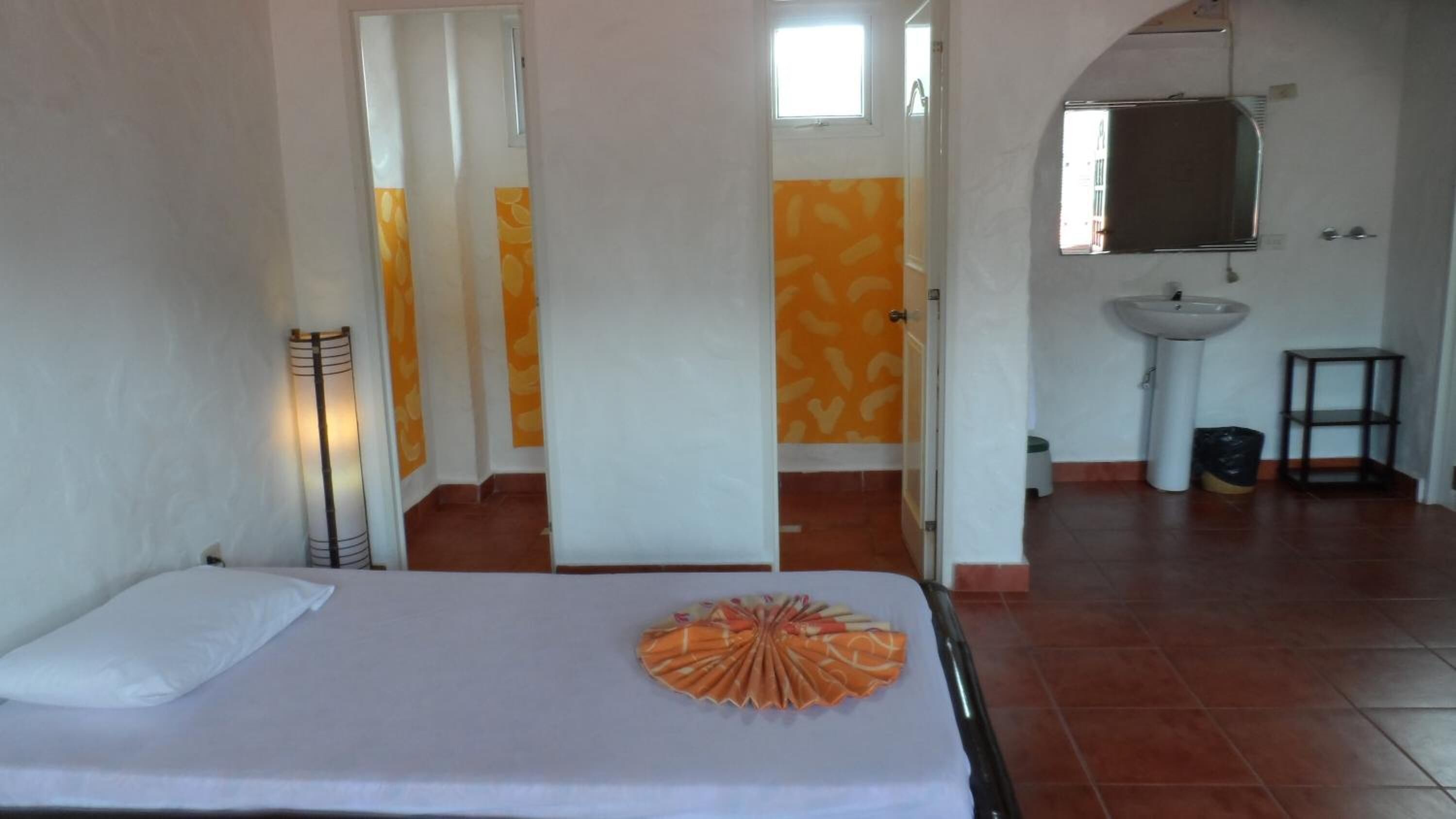 Ein Hotelzimmer mit Bett, Nachttischlampe, einem dekorativen orangefarbenen Fächer, einem Badezimmer mit Waschbecken und Spiegel sowie einem gefliesten Boden.