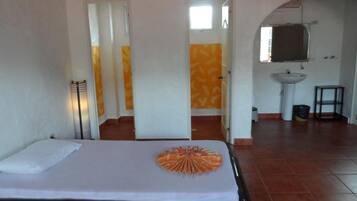 Una habitación de hotel con cama, lámpara de noche, un abanico decorativo anaranjado, baño con lavamanos y espejo, y suelo de baldosas.