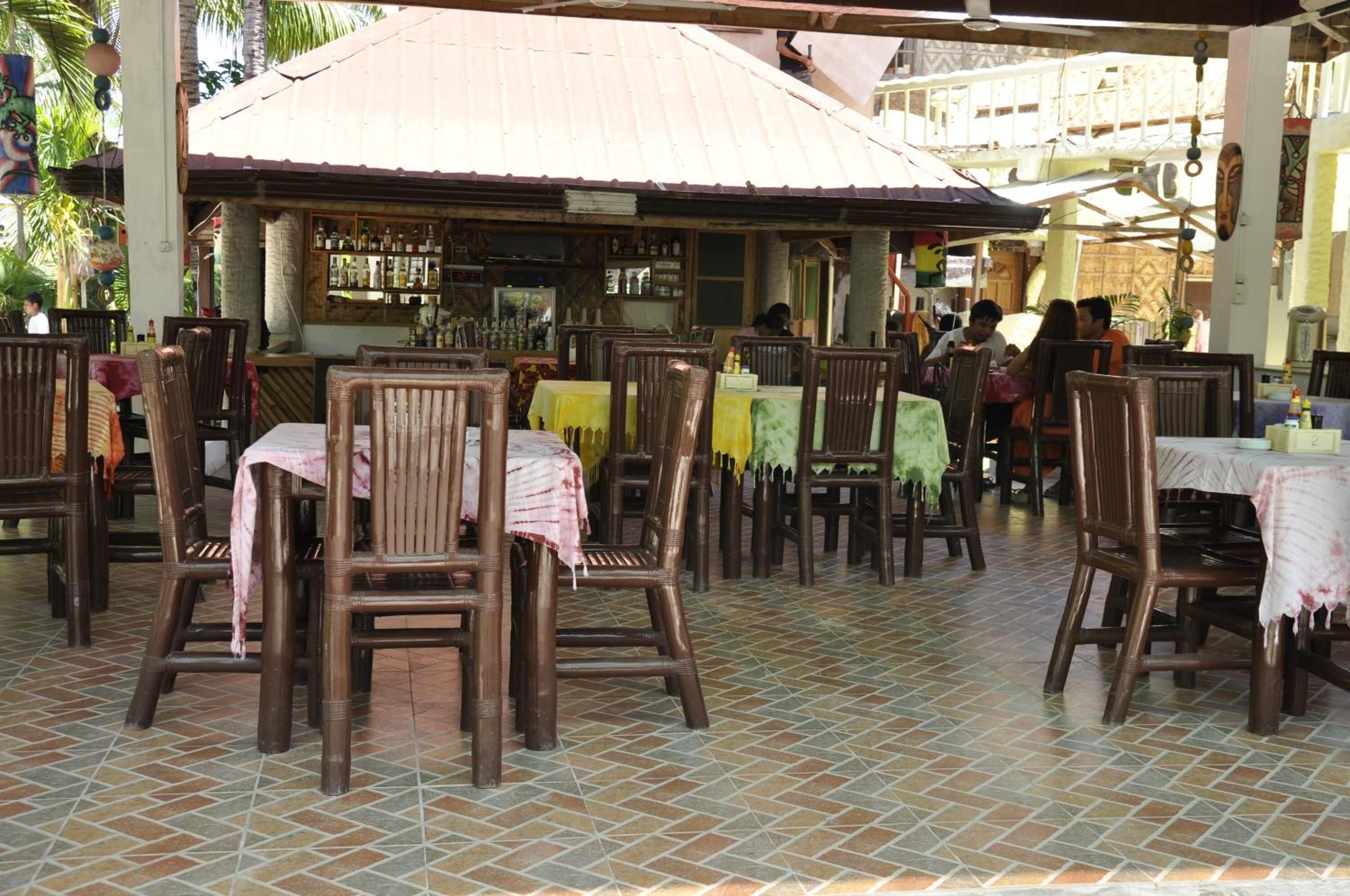 Un restaurante al aire libre con mesas y sillas de madera, suelo de baldosas y una zona de bar al fondo.