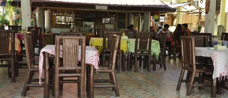 Un restaurante al aire libre con mesas y sillas de madera, suelo de baldosas y una zona de bar al fondo.