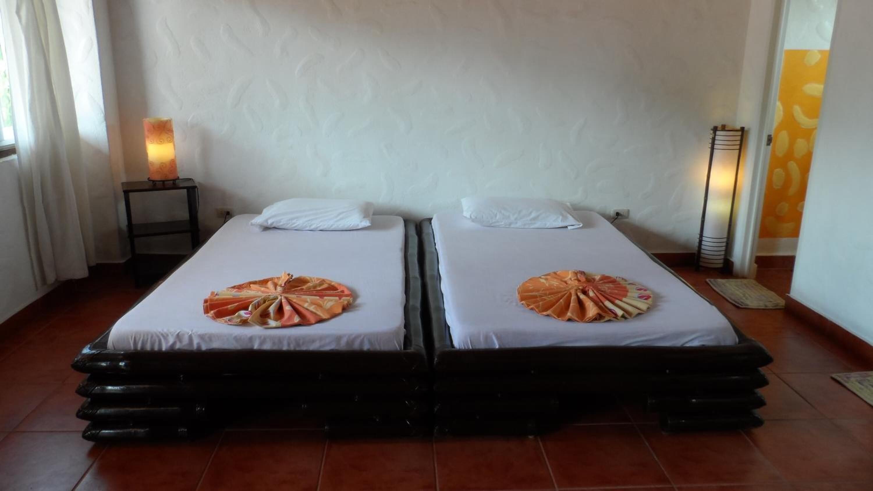 Une chambre avec deux lits, sur chacun desquels une serviette est pliée en forme d’éventail. Une table de chevet avec une lampe se trouve entre les deux lits, et un radiateur fixé au mur est placé sur le côté droit.