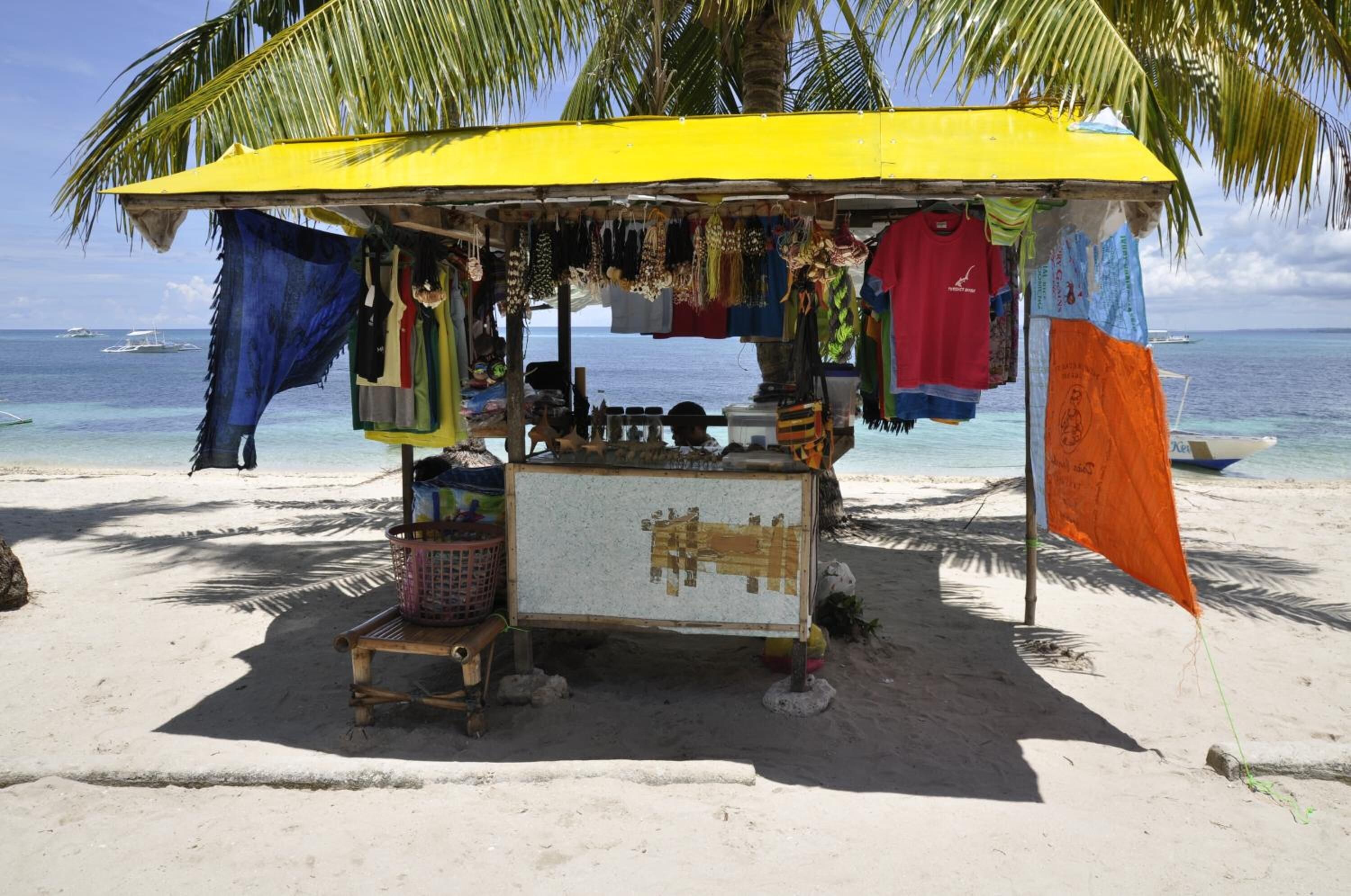 A beachside market stall with various items hanging for sale, a small table with goods, and a clear view of the ocean and boats in the background.