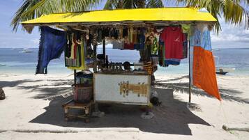 A beachside market stall with various items hanging for sale, a small table with goods, and a clear view of the ocean and boats in the background.