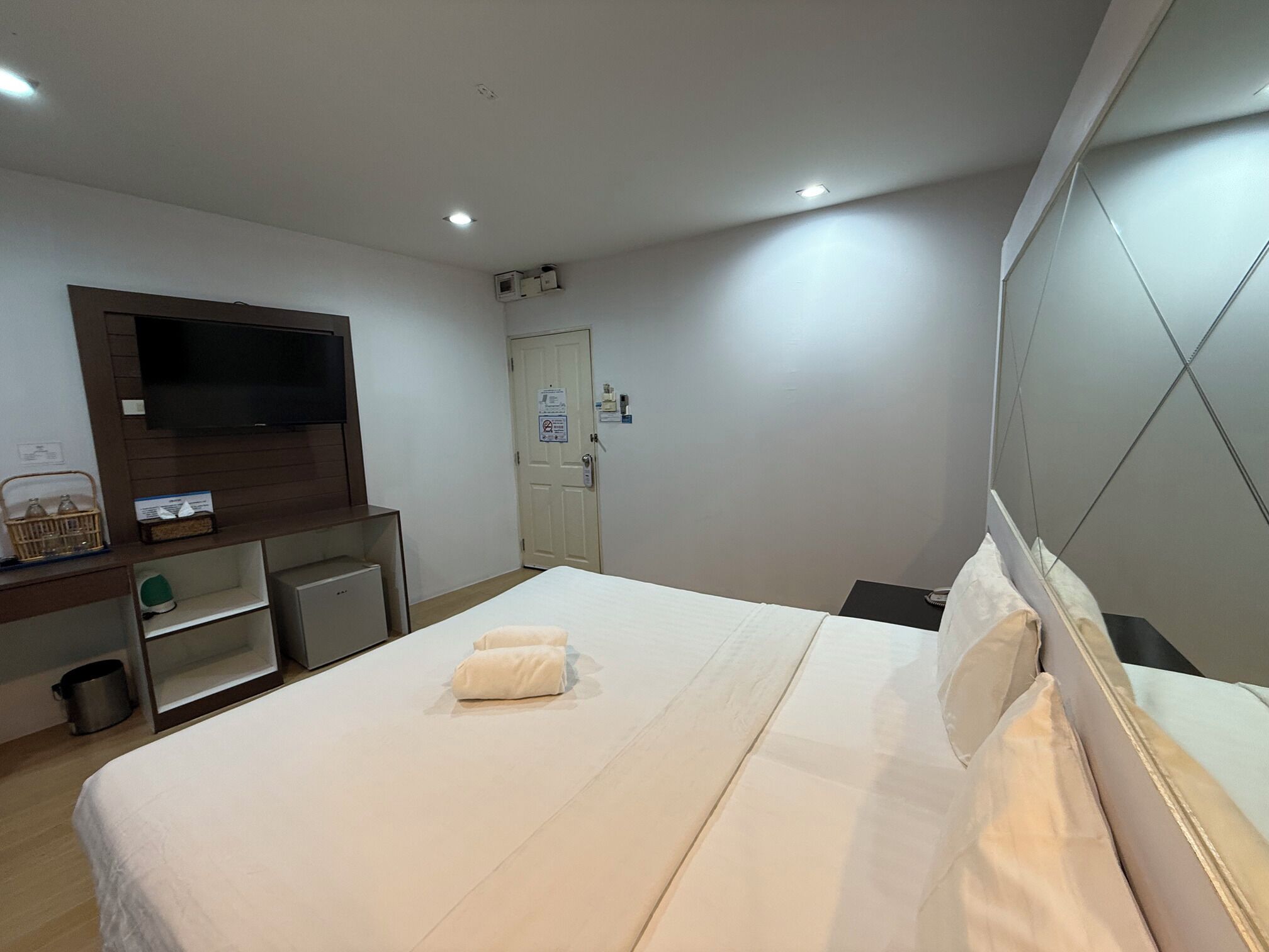 A hotel room with a bed, a TV mounted on the wall, a desk with a computer, and a door with a sign.