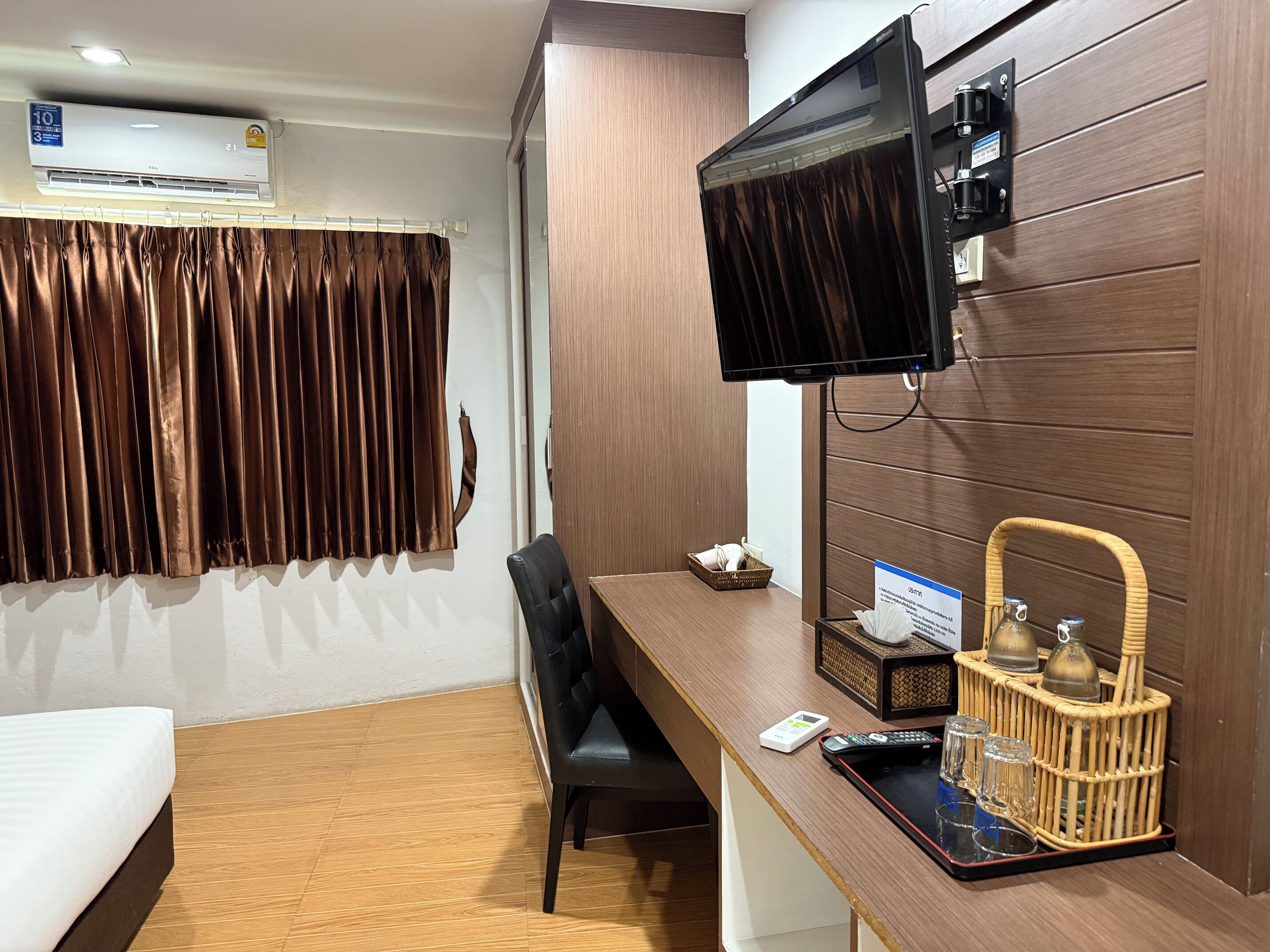 A hotel room with a wooden desk, a television mounted on the wall, a bed with a white mattress, and a window with brown curtains.