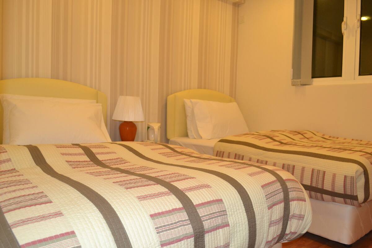 A hotel room with two beds, each with a striped bedspread, a bedside table with a lamp, and a window with blinds.