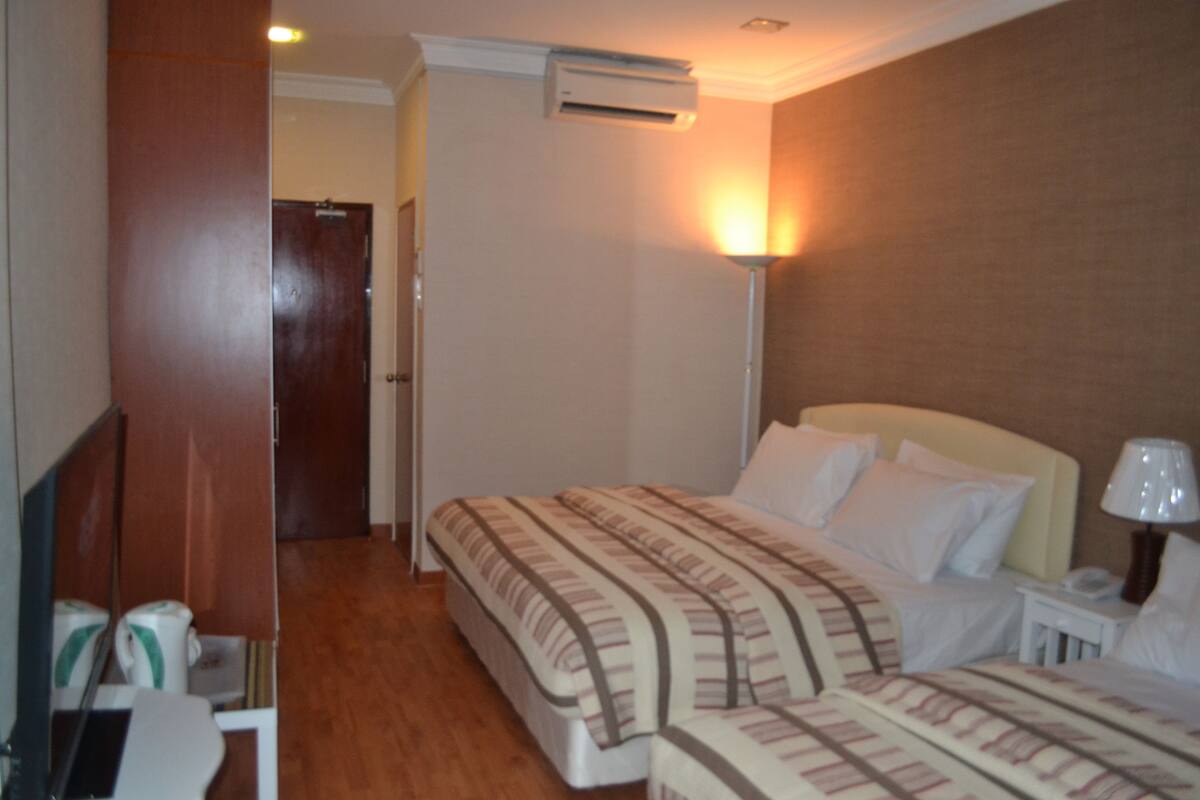 A hotel room with two beds, a bedside table with a lamp, an air conditioning unit, and a wooden floor.