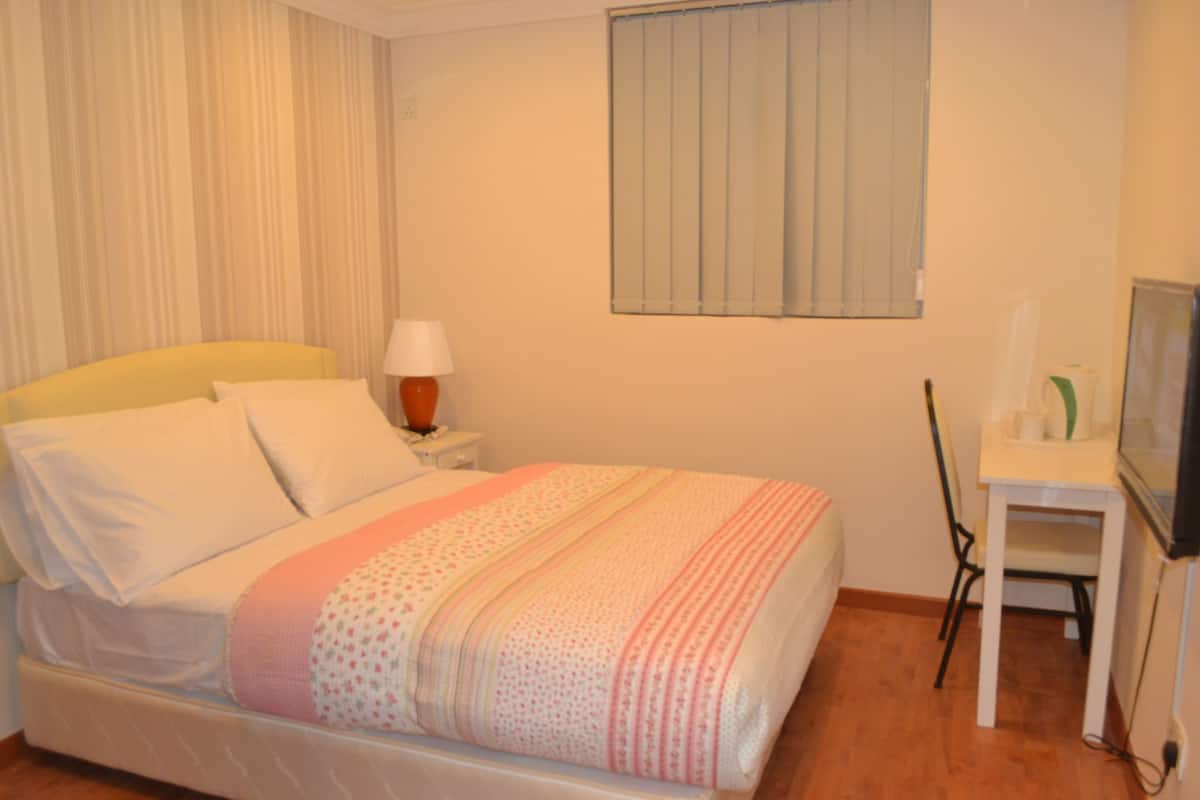 A neatly made bed with a striped blanket, a bedside table with a lamp, a chair, and a television on a stand.