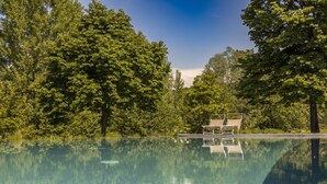 A clear swimming pool surrounded by lush trees, with a small structure and two lounge chairs in the background.
