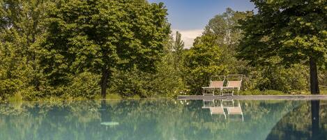 A clear swimming pool surrounded by lush trees, with a small structure and two lounge chairs in the background.
