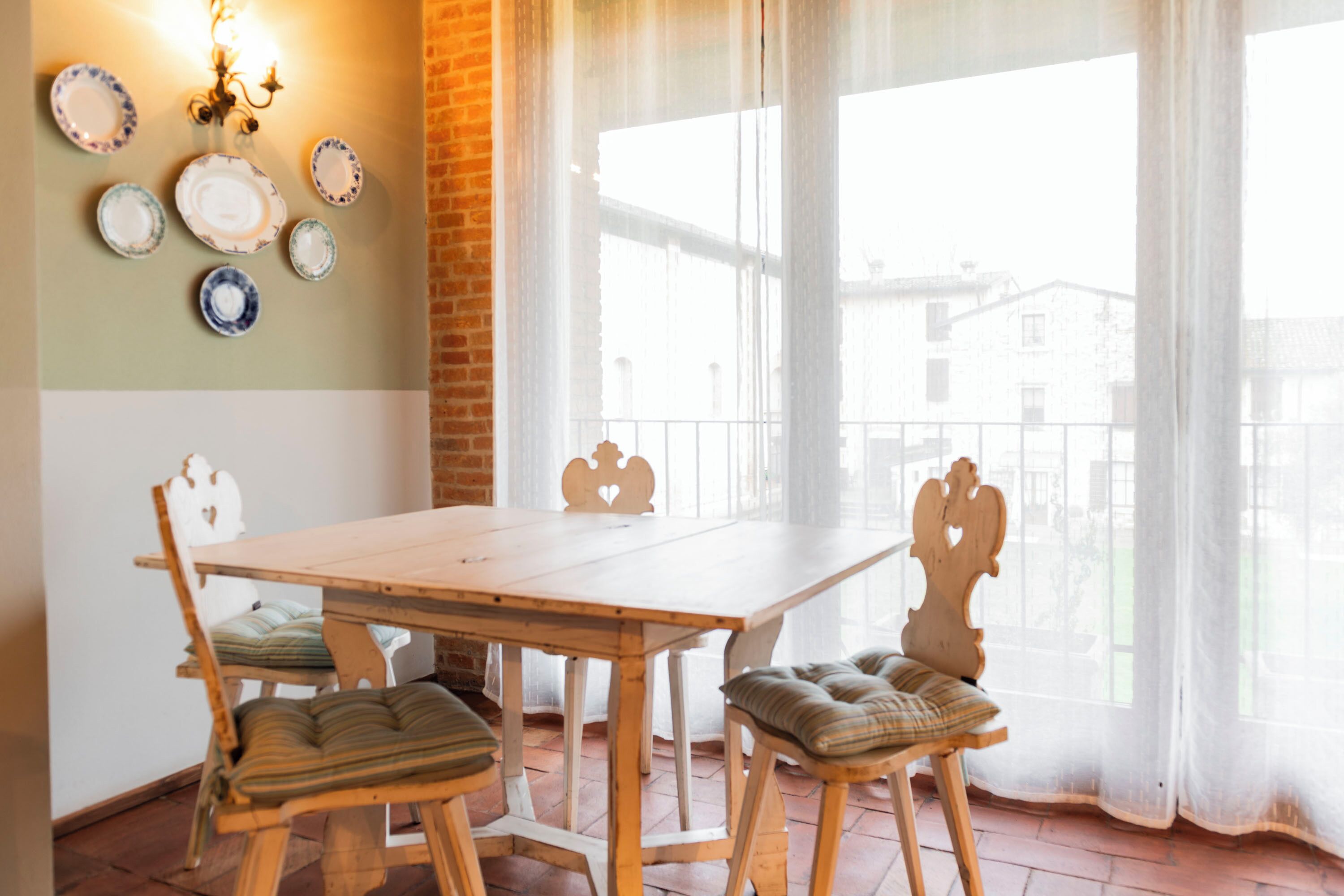 A dining area with a wooden table and chairs, a brick wall, and a view through large windows.