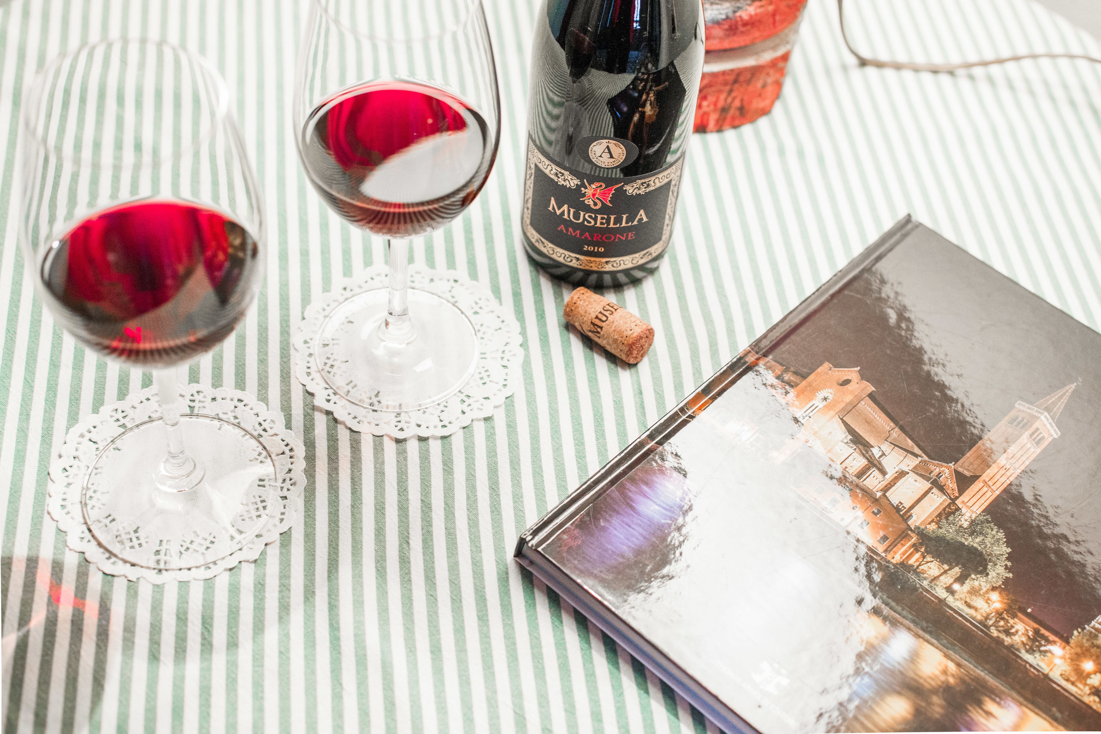 Una bottiglia di vino Amarone Musella, due bicchieri da vino, un tappo e un libro con un'immagine di un panorama urbano.