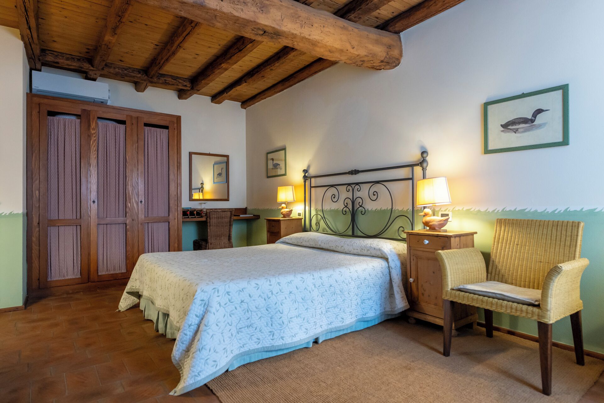 Een slaapkamer met een houten plafond, een bed met een beddegoed met een patroon, een rieten stoel, een nachtkastje met een lamp en een aan de muur bevestigd schilderij.