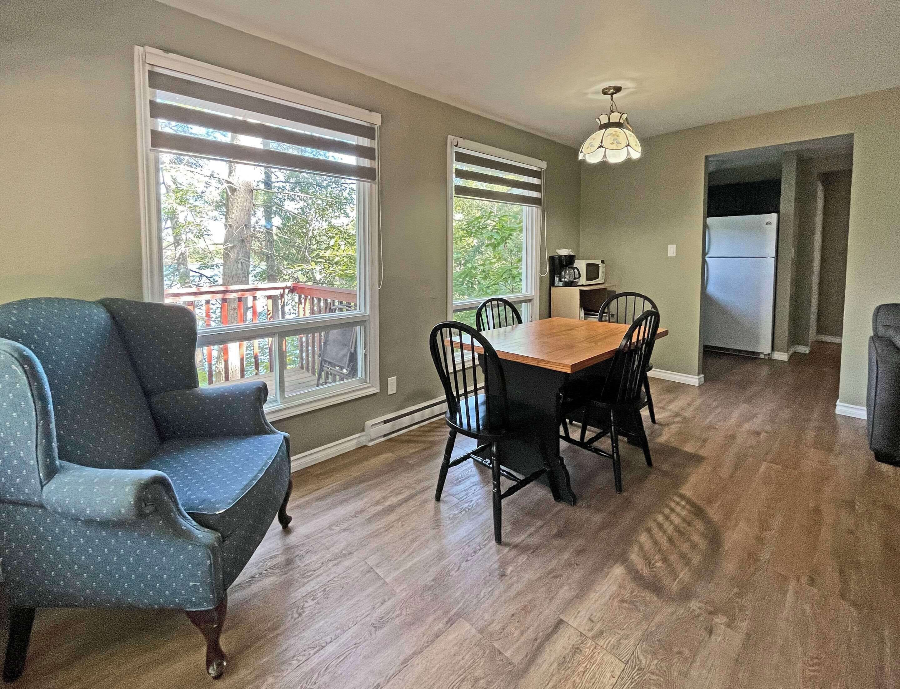 A dining area with a wooden table and chairs, a blue armchair, and large windows with blinds.