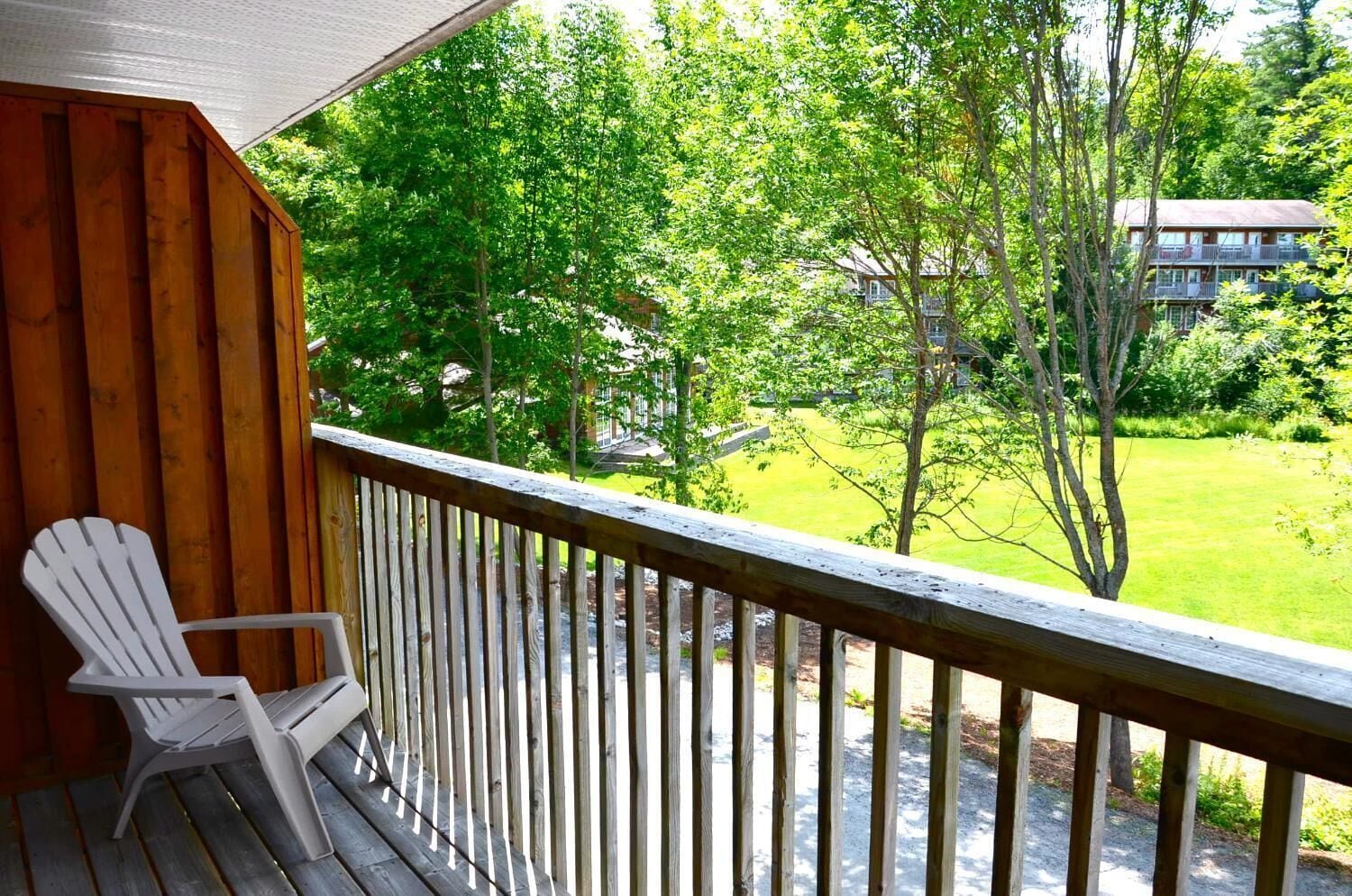 A wooden deck with a white Adirondack chair, overlooking a grassy area with trees and a building in the background.