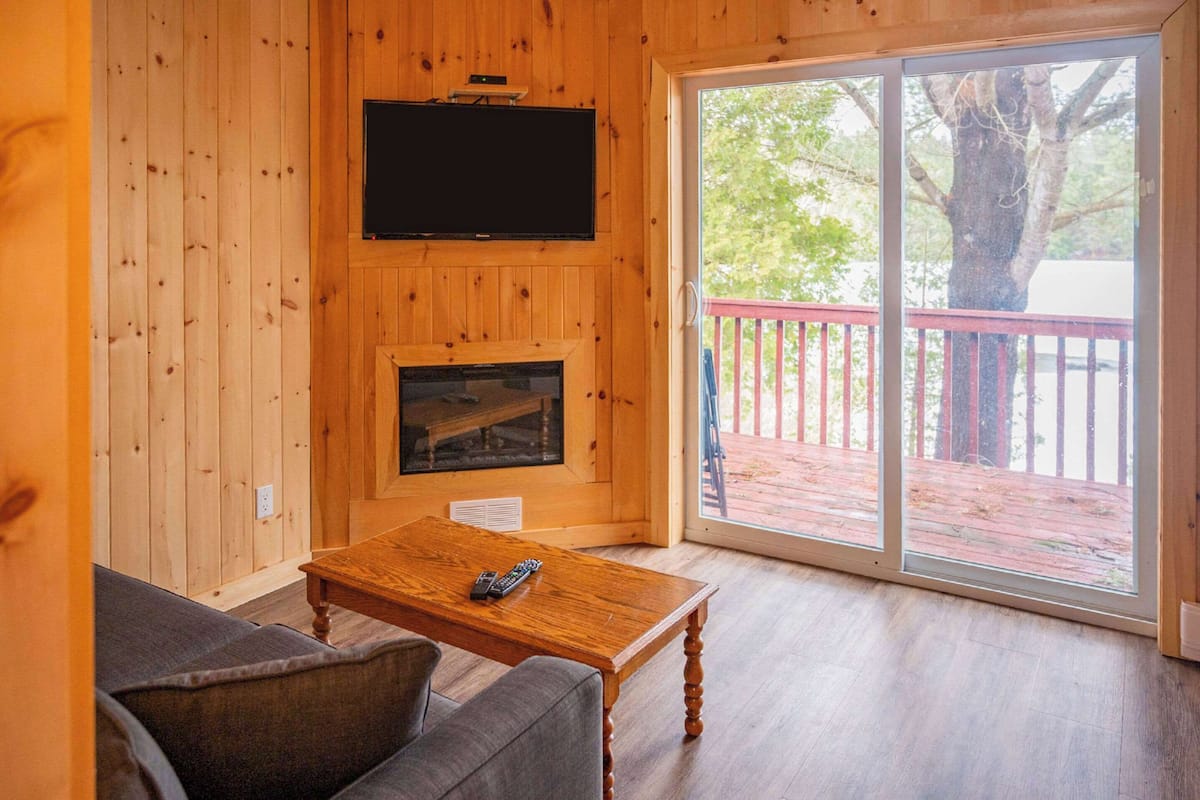 Ein Zimmer mit Holzwandverkleidung, einem Kamin, einem darüber montierten Fernseher, einer Couch und einer Glastür zum Außenbereich.