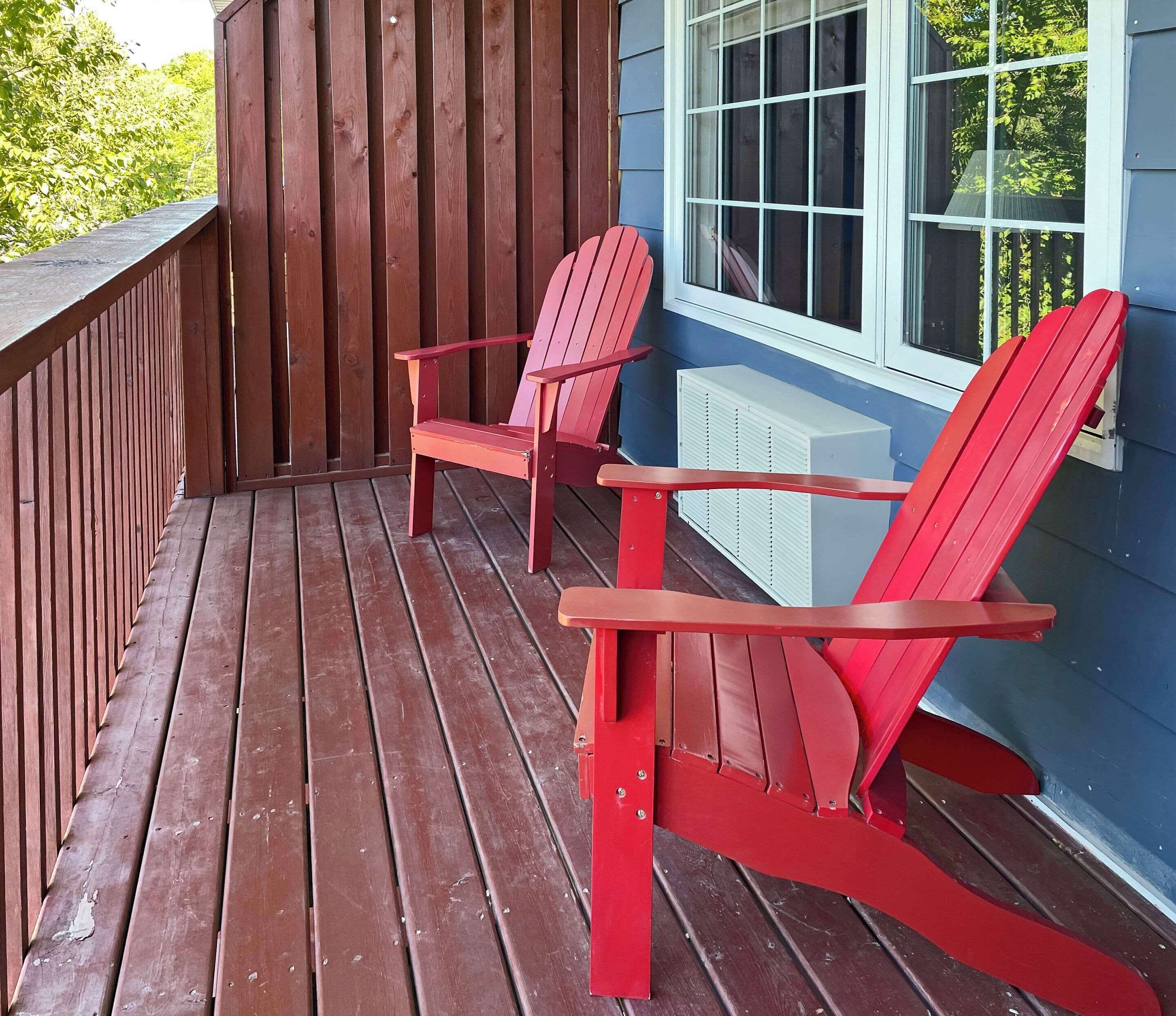 Una terraza de madera con dos sillas Adirondack rojas y una unidad de aire acondicionado blanca.