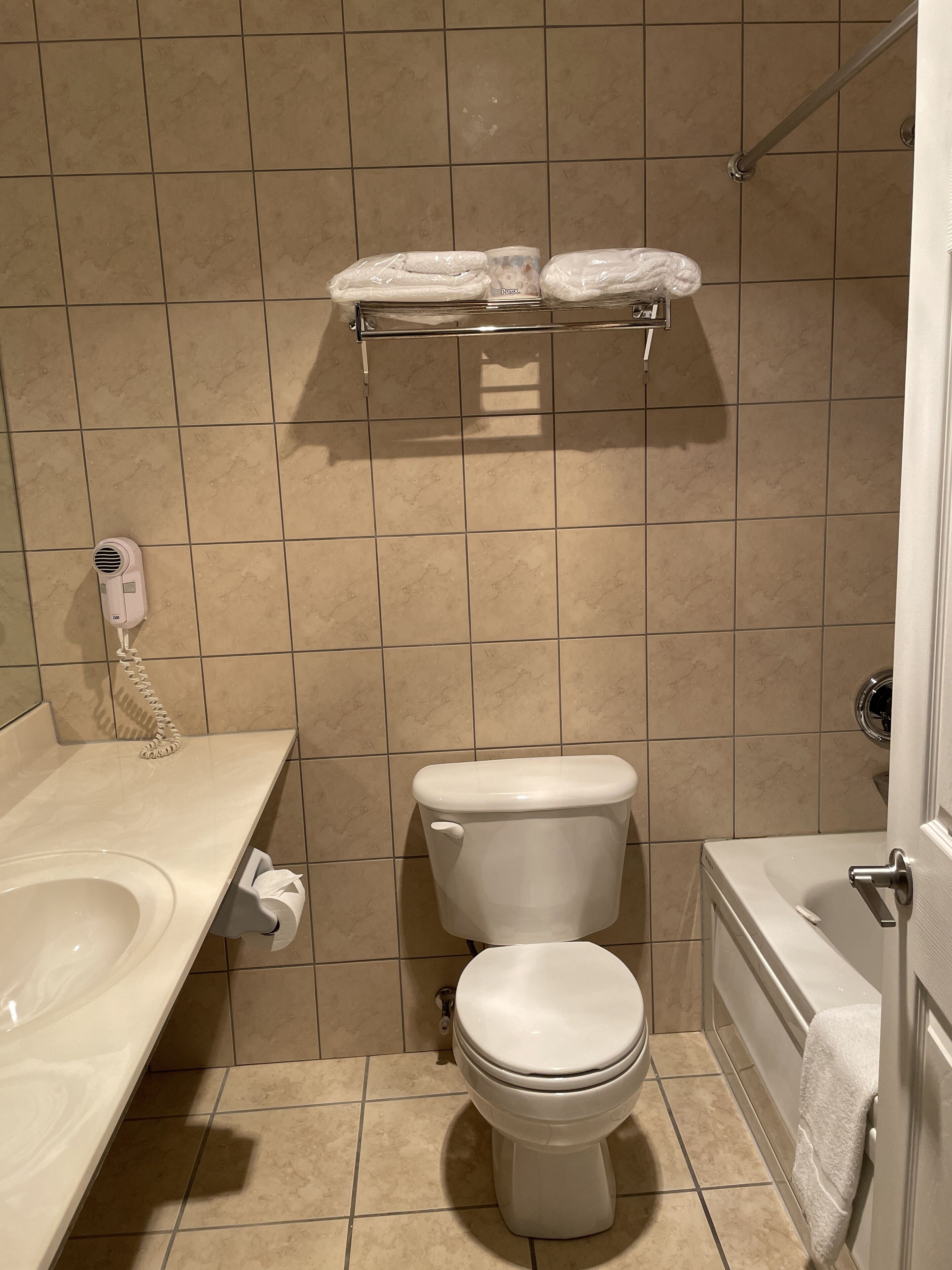 Un baño pequeño con inodoro, lavamanos y bañera. Hay un perchero sobre el inodoro y un secamanos en la encimera.