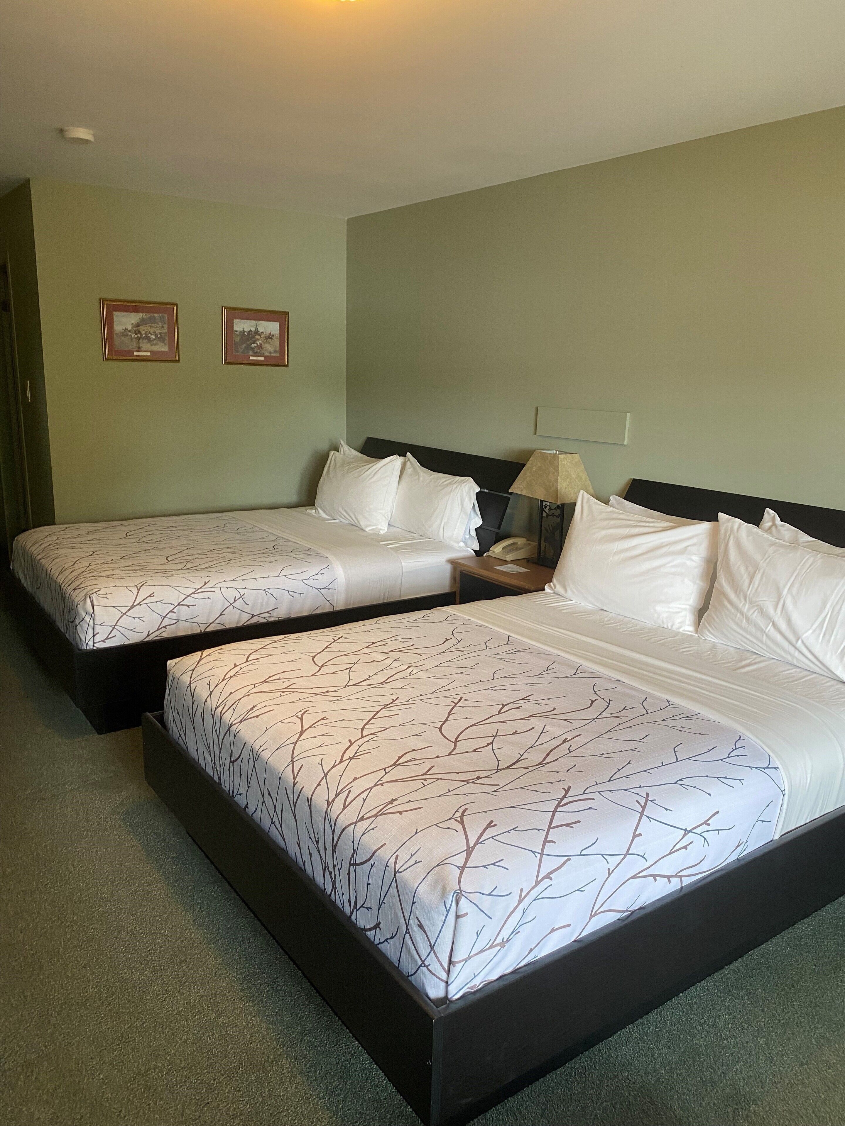 Quarto de hotel com duas camas de solteiro, uma mesinha de cabeceira com abajur e duas imagens emolduradas na parede.