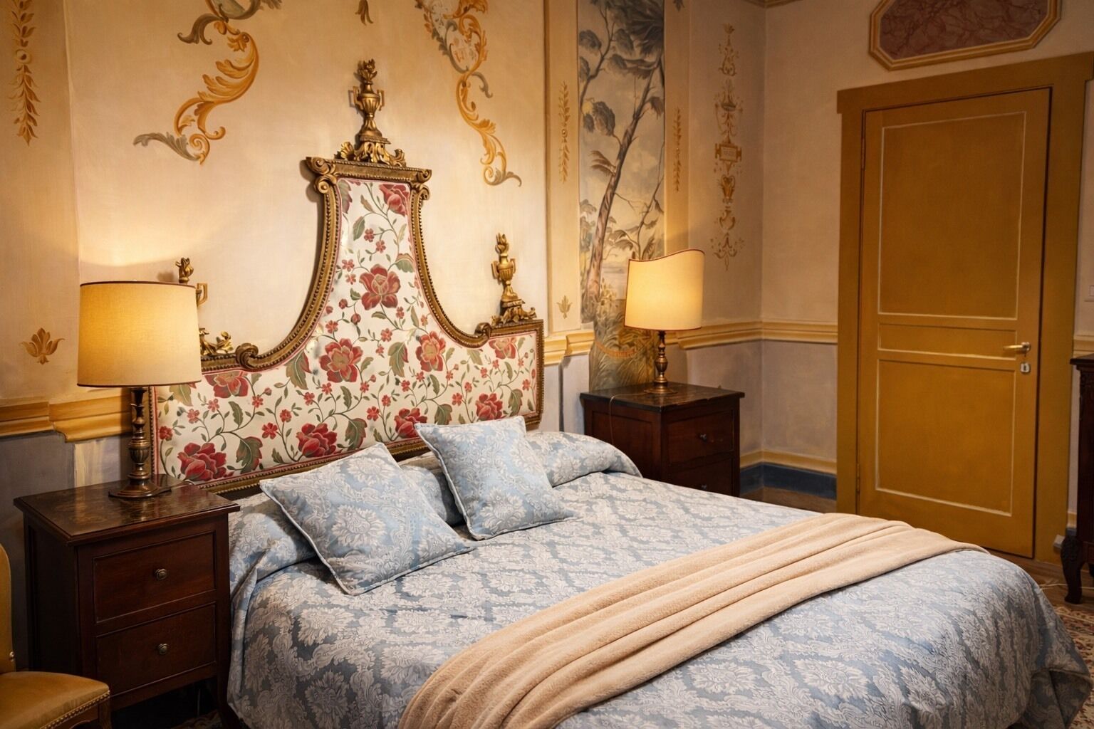 Un dormitorio clásico con un cabecero floral, dos mesitas de noche con lámparas, una cama grande con colcha estampada azul y un armario de madera.