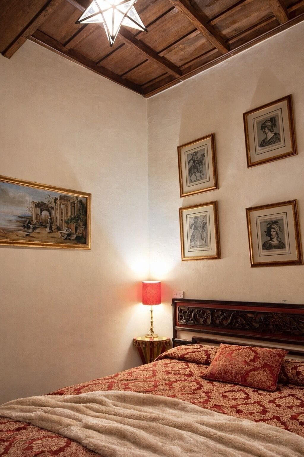 Un dormitorio con techo de madera, una cama con ropa de cama rojo y beige, una mesita de noche con una lámpara roja y tres cuadros en la pared.