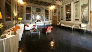 Una habitación amplia con techo alto, vigas ornamentales y suelo negro pulido. Hay varias zonas de estar con sofás y sillones tapizados en blanco y rojo. Las paredes están decoradas con cuadros enmarcados y paneles decorativos.